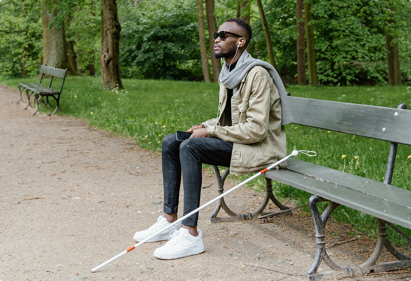 a blind man sitting and listening to a phone, with a white cane propped next to him