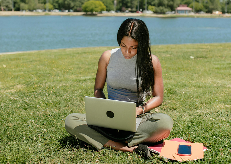 Woman working on a laptop on a grassy lawn in front of a river